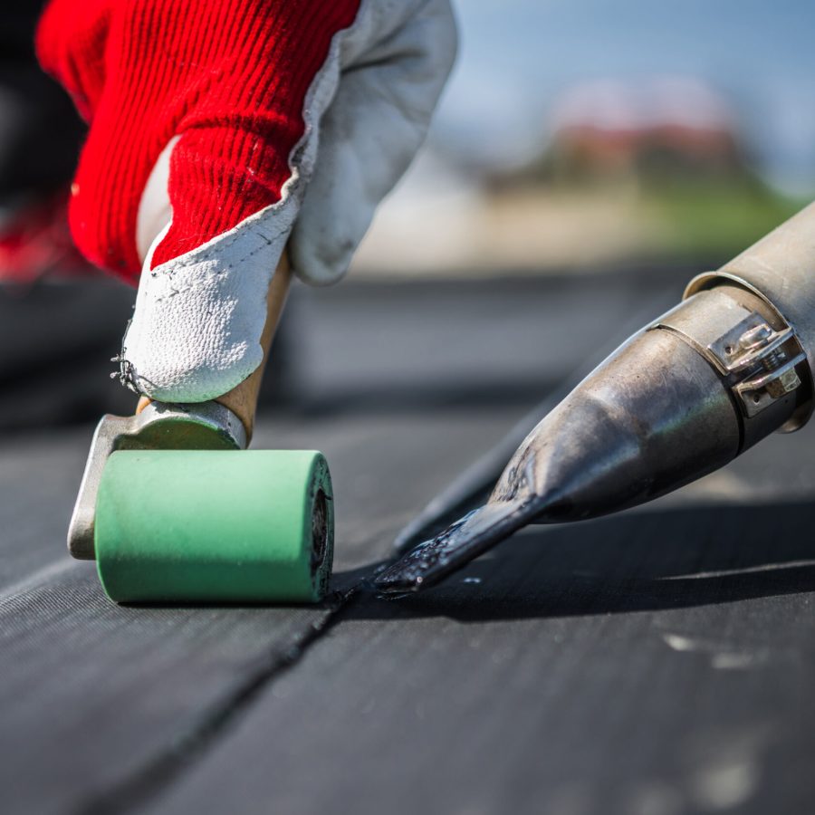 Roof Worker Attaching Pieces of EPDM Membrane Roofing Material Using Hot Air Blower and Industrial Roller.